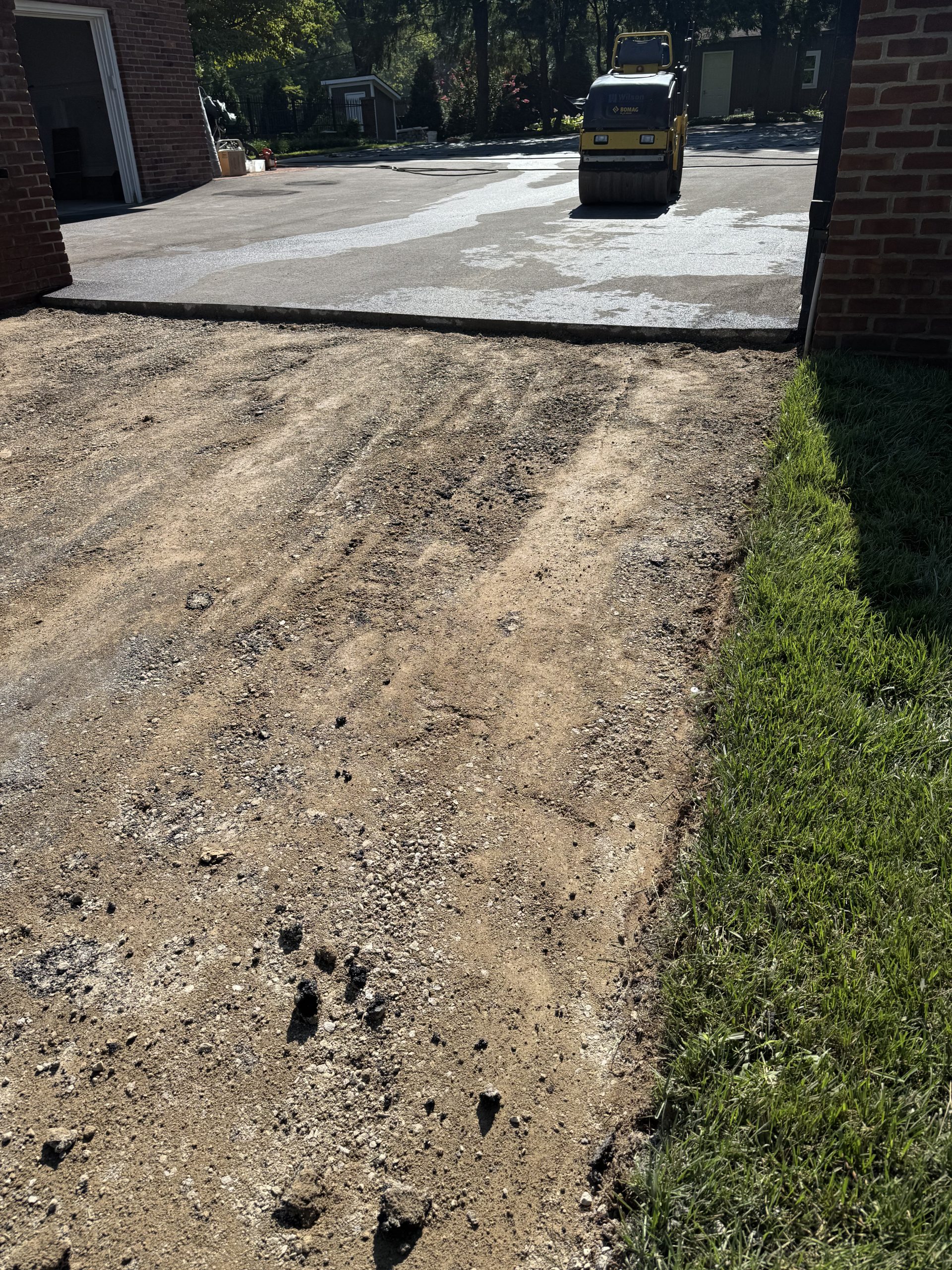 Wide view of the excavated driveway section prepped for repair, with a paving roller compacting freshly laid asphalt